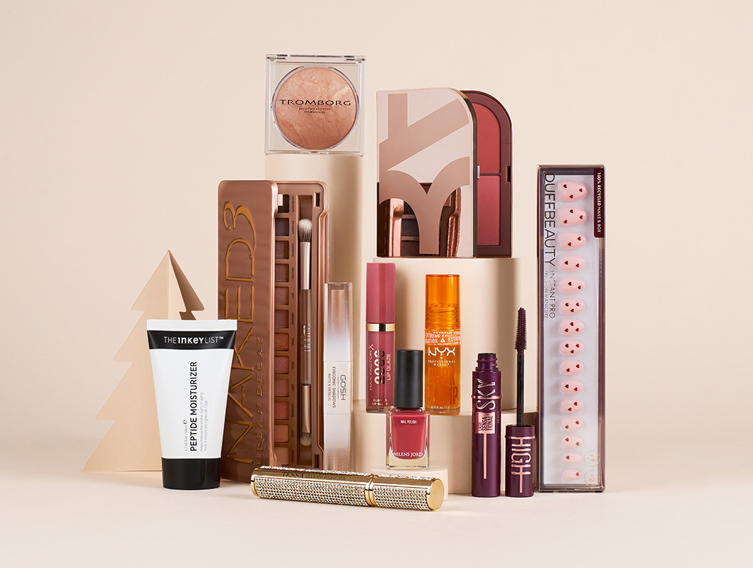 Udvalg af makeup- og negleprodukter fra blandt andet Tromborg, Urban Decay, GOSH, NYX og DUFFBEAUTY står opstillet på lyse podier med dekorative juletræer i beige nuancer.