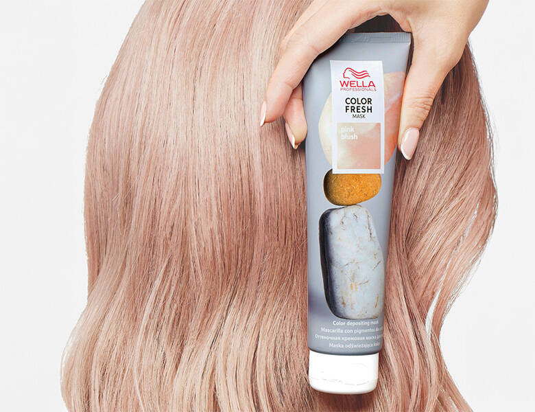 Her ses et billede af en person med lyst hår, der holder et hårprodukt fra Wella Professionals op mod håret. På tuben står teksten “COLOR FRESH MASK pink blush”. Produktet er i lyse nuancer med pink detaljer.