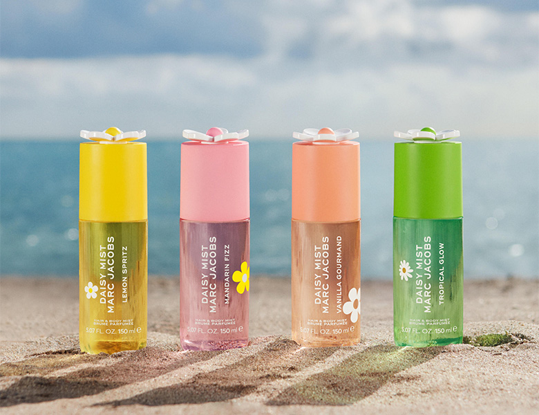 Her ses et billede med fire Daisy Mist Hair & Body Mist-produkter fra Marc Jacobs placeret på sand med havet i baggrunden. Produkterne vises i varianterne Lemon Spritz, Mandarin Fizz, Vanilla Gourmand og Tropical Glow.
