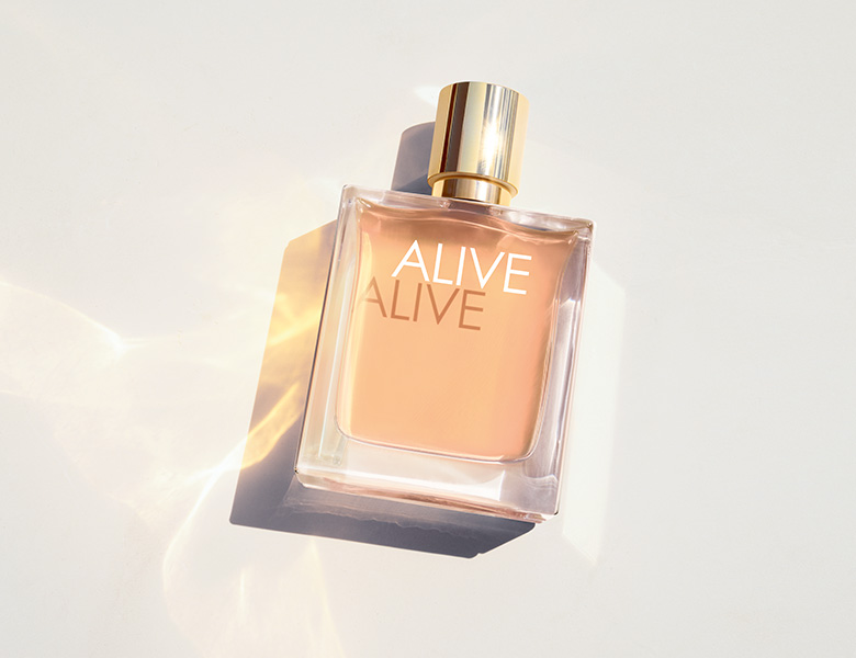 Her ses et billede med en parfumeflakon fra BOSS Alive placeret mod en lys baggrund. På flakonen står teksten “ALIVE”, og den gyldne kork og lyse rosatoner fremhæves af lyse refleksioner omkring produktet.