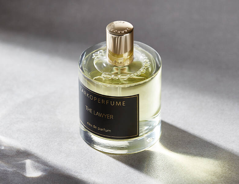 Her ses et billede af en parfumeflakon fra ZARKOPERFUME placeret på en lys overflade. På flakonen står teksten “ZARKOPERFUME THE LAWYER eau de parfum”. Flakonen er rund og gennemsigtig med gyldne nuancer og har et guldfarvet låg.