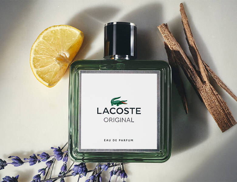 Her ses en lys baggrund med nogle blomster, kanel og lime. I midten ligger en grøn og hvid Lacoste parfume.