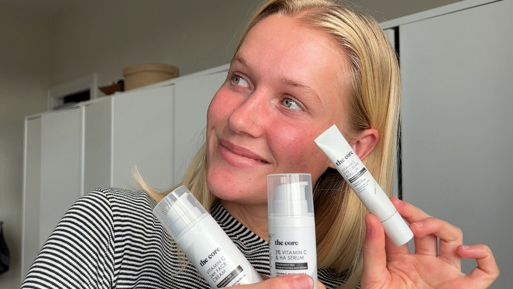En smilende person med lyst hår holder tre hudplejeprodukter fra serien “The Core” op foran sig. Personen ser væk fra kameraet og er iført en sort- og hvidstribet trøje. Produkterne består af en ansigtscreme, et serum og en øjencreme i hvide beholdere med sort tekst. Billedet er taget indendørs foran hvide skabe og har et naturligt, hverdagsagtigt udtryk.