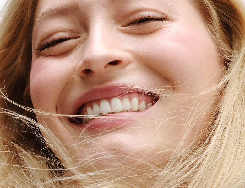 Her ses et nærbillede af en kvinde, der smiler med lukkede øjne. Ansigtet fylder billedet, og håret bevæger sig let foran ansigtet.