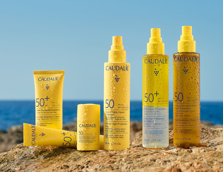 Her ses CAUDALIE solprodukter i gule flasker placeret på klipper ved havet.