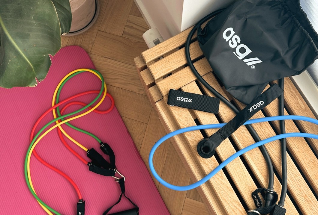 Farverige træningselastikker og andet træningstilbehør ligger ved en pink træningsmåtte og en træbænk fra ASG