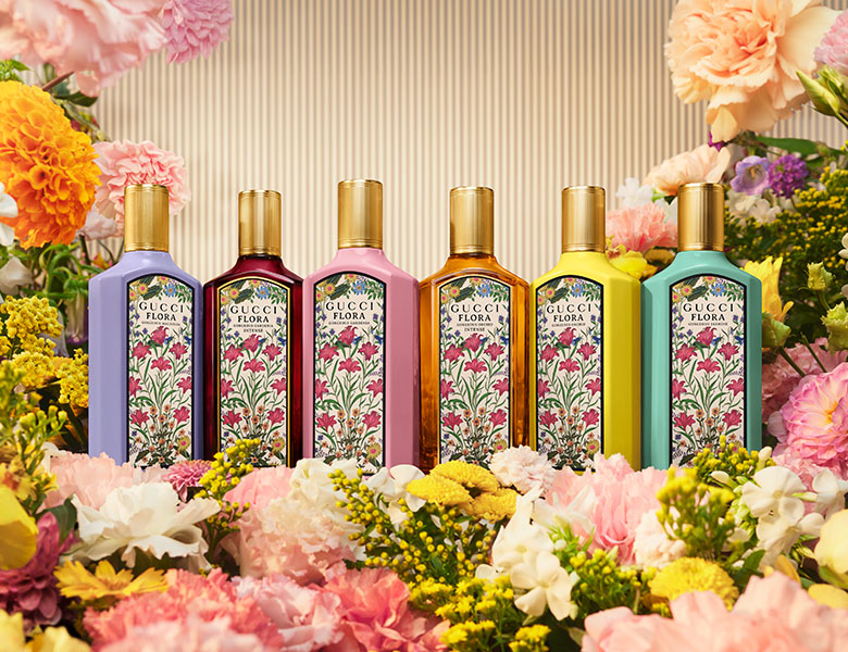 Her ses et billede med seks parfumeflakoner fra Gucci Flora i forskellige farver placeret blandt farverige blomster mod en lys baggrund. Flakonerne har blomstrede etiketter og gyldne låg og repræsenterer forskellige varianter i Gucci Flora-serien.