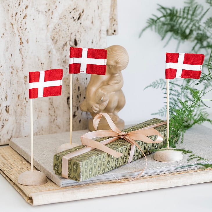 Tre små danske flag står omkring en aflang gave, der er pakket ind i pengeseddelmønster og bundet med et lyserødt bånd, med grønne grene og en dekorativ figur i baggrunden.