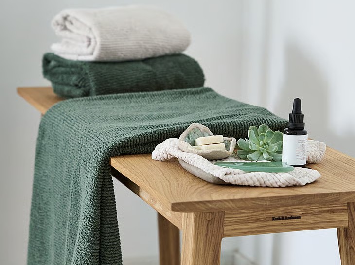 Grønne og lyse håndklæder ligger foldet på en træbænk sammen med små wellnessprodukter, sæbe og en sukkulent i rolige, naturlige omgivelser.