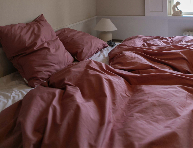 Her ses en seng redt med rosa sengetøj og puder i et lyst soveværelse.