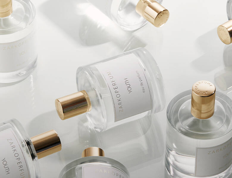 Her ses et billede med flere parfumeflakoner fra ZARKOPERFUME placeret spredt på en lys, reflekterende overflade. På flakonerne står teksten “ZARKOPERFUME YOUTH eau de parfum”. Flakonerne er gennemsigtige med klare væsker og har guldfarvede låg.