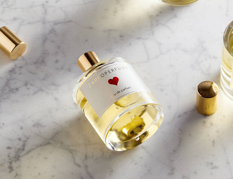 Her ses et billede af en parfumeflakon fra ZARKOPERFUME placeret på en lys marmorlignende overflade. På flakonen står teksten “ZARKOPERFUME” og “eau de parfum”, og etiketten har et lille rødt hjerte. Flakonen er gennemsigtig med gyldne nuancer og har et guldfarvet låg.