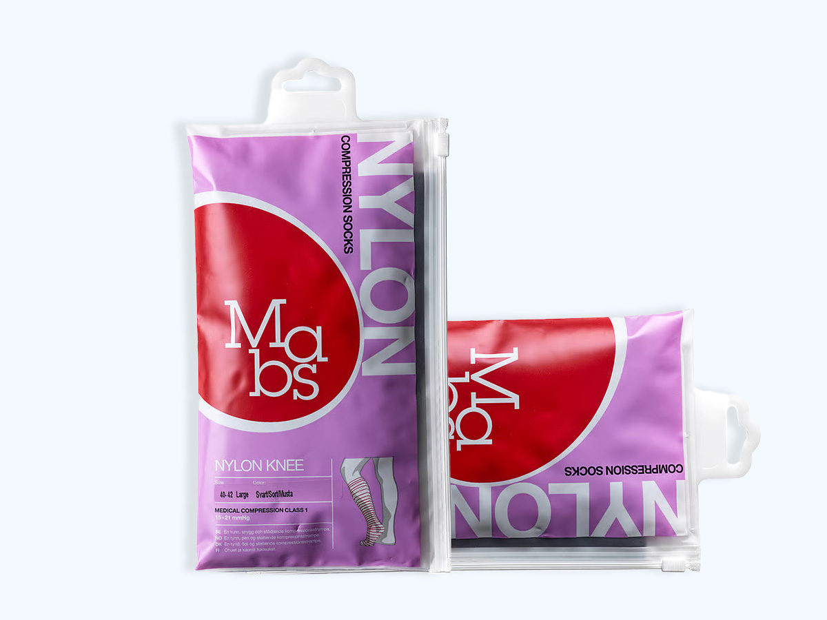 Her ses et billede med to pakker kompressionsstrømper fra Mabs mod en lys baggrund. Produkterne ligger i gennemsigtige genlukkelige poser med lilla og rødt design. På emballagen står blandt andet “Compression Socks”, “Nylon Knee” og “Medical Compression Class 1”, og den ene pakke viser en illustration af knæstrømperne.