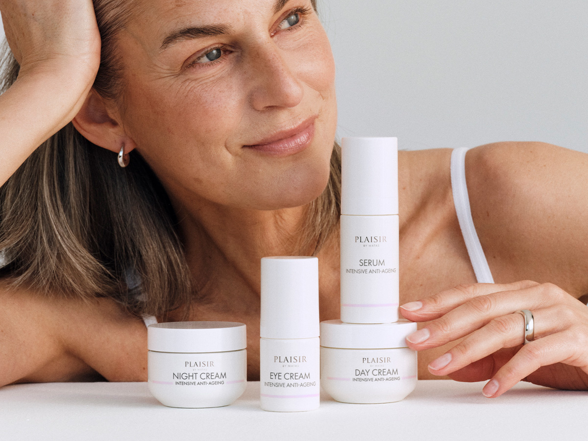 Her ses en kvinde med kort, lyst hår og hvid top, der læner sig op ad fire Plaisir hudplejeprodukter: natcreme, øjencreme, dagcreme og serum. Baggrunden er lys og ensfarvet med blødt dagslys, som giver et roligt og eksklusivt udtryk.