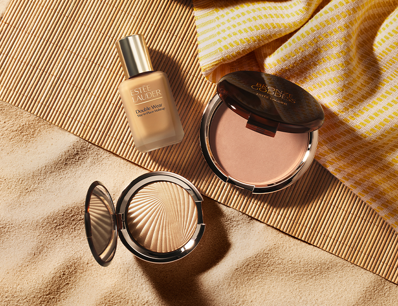 Her ses et billede med makeup-produkter fra Estée Lauder placeret i sand. Der ses blandt andet foundationen “Double Wear” samt bronzer- og pudderprodukter i kompakter.