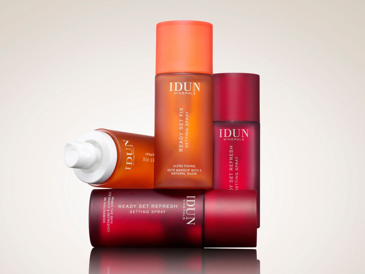 Her ses et billede med setting sprays fra IDUN Minerals mod en lys baggrund. Produkterne omfatter Ready Set Fix Setting Spray og Ready Set Refresh Setting Spray, arrangeret i røde og orange flasker med matte overflader. Produkterne er stablet, så der skabes fokus på de forskellige varianter.