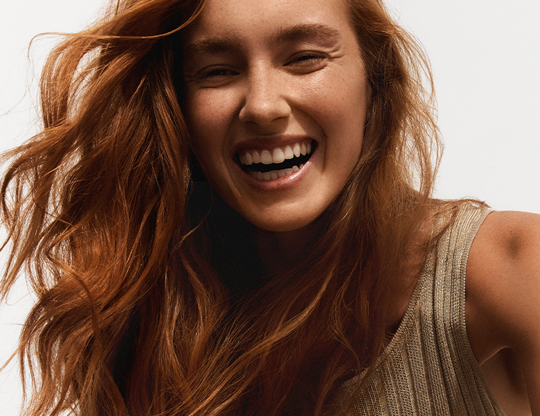 Her ses en smilende person med langt, bølget rødt hår iført en beige top mod en lys baggrund.