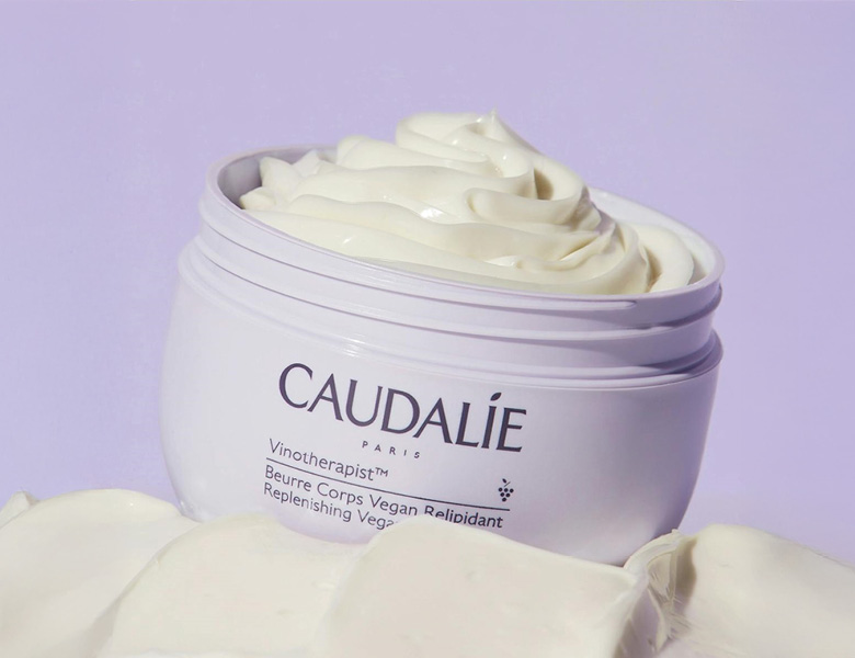 Her ses en CAUDALIE body cream i en krukke med creme på en lys lilla baggrund.