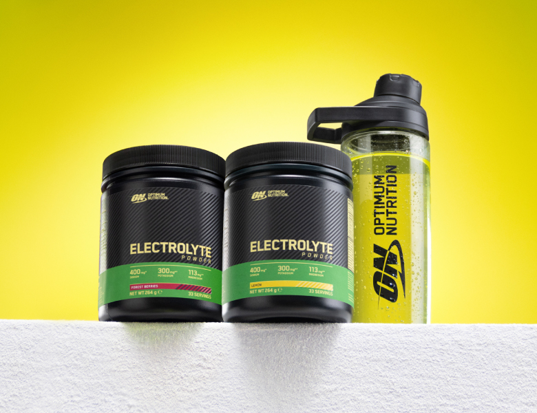 To bøtter Optimum Nutrition Electrolyte Powder og en gennemsigtig drikkedunk med gul væske står på et hvidt håndklæde foran en gul baggrund.