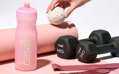 Pink træningsmåtte, drikkeflaske, håndvægte, elastikker og massagebold fra Casall på lyserød og hvid baggrund.
