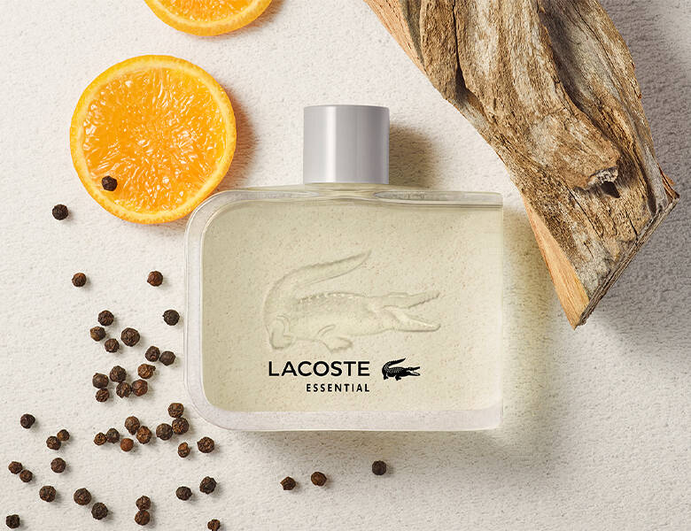 Her ses en lys baggrund med appelsin, træ og peberkorn. I midten ligger en lys Lacoste parfume.