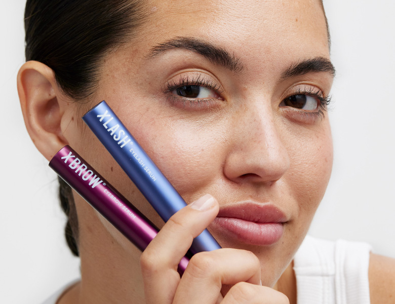 Her ses en person, der holder Xlash vippeserum og Xbrow brynserum foran ansigtet.
