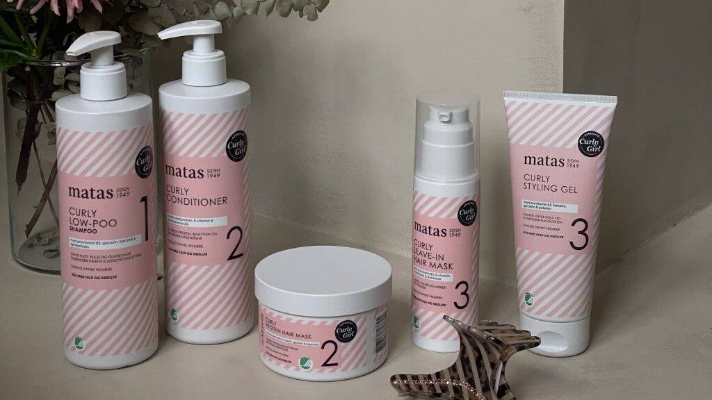 Flere hårprodukter fra Matas Curly-serien står opstillet på en lys overflade indendørs. Produkterne har hvide emballager med lyserøde striber og består blandt andet af shampoo, conditioner, leave-in hårmaske, stylinggel og en rund hårmaske i krukke. Flaskerne står side om side, mens krukken er placeret foran dem. Forrest i billedet ligger en mørk hårklemme. I baggrunden ses tørrede blomster, som giver billedet en blød og hyggelig stemning.