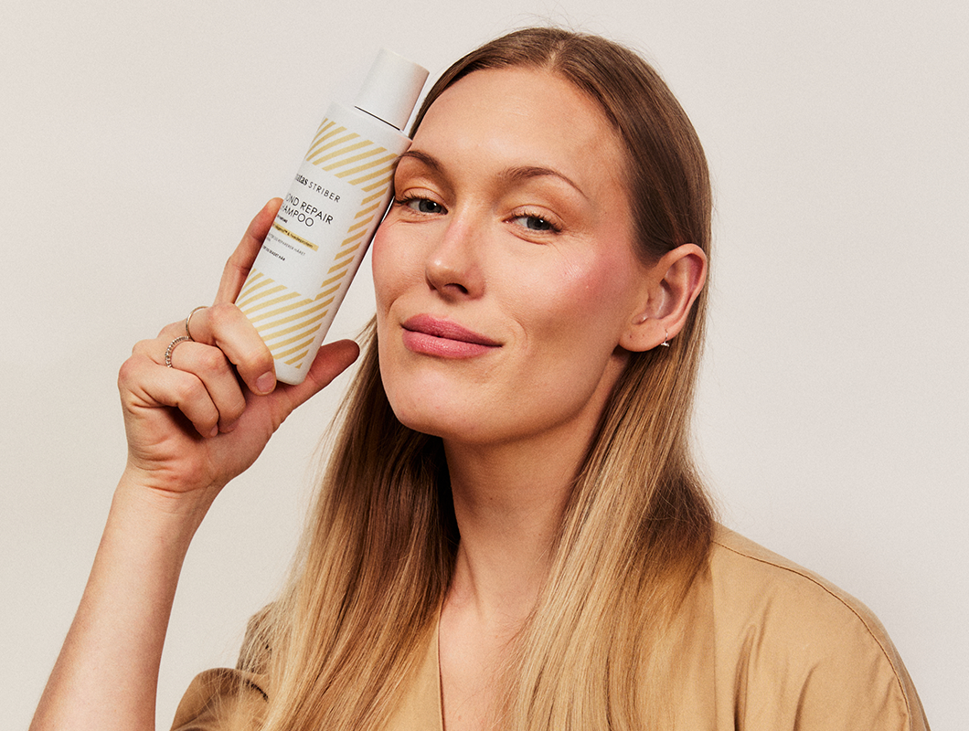 Her ses et nærbillede af en person, der holder en flaske Matas Striber Bond Repair Shampoo op ved ansigtet. Personen ser direkte mod kameraet med et let smil, mens produktet holdes tæt ved kinden. Baggrunden er lys og enkel, hvilket giver fokus til personen og shampooen.