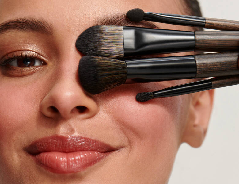 Her ses en kvindes ansigt. Hun holder makeupbørster over det ene øje.
