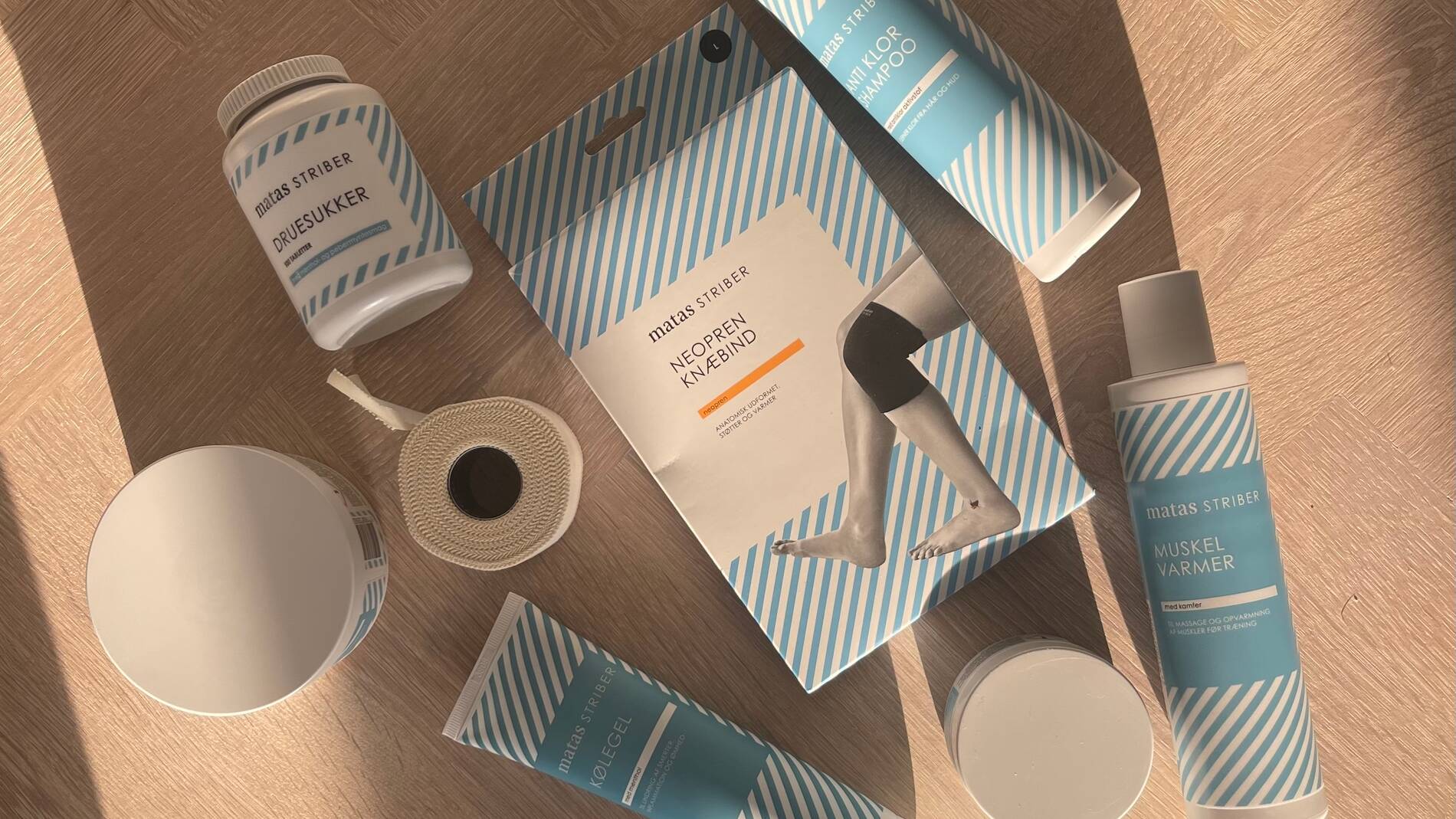 Flere produkter fra Matas Striber ligger arrangeret på et lyst trægulv set ovenfra i varmt sollys. Produkterne har hvide emballager med lyseblå striber og består blandt andet af anti-klø shampoo, kølegel, muskelvarmer, druesukker og et neopren-knæbind i emballage med billede af et knæ. Der ligger også en rulle sportstape samt flere runde krukker mellem produkterne. Sollyset skaber lange skygger hen over gulvet og giver billedet en varm og rolig stemning.