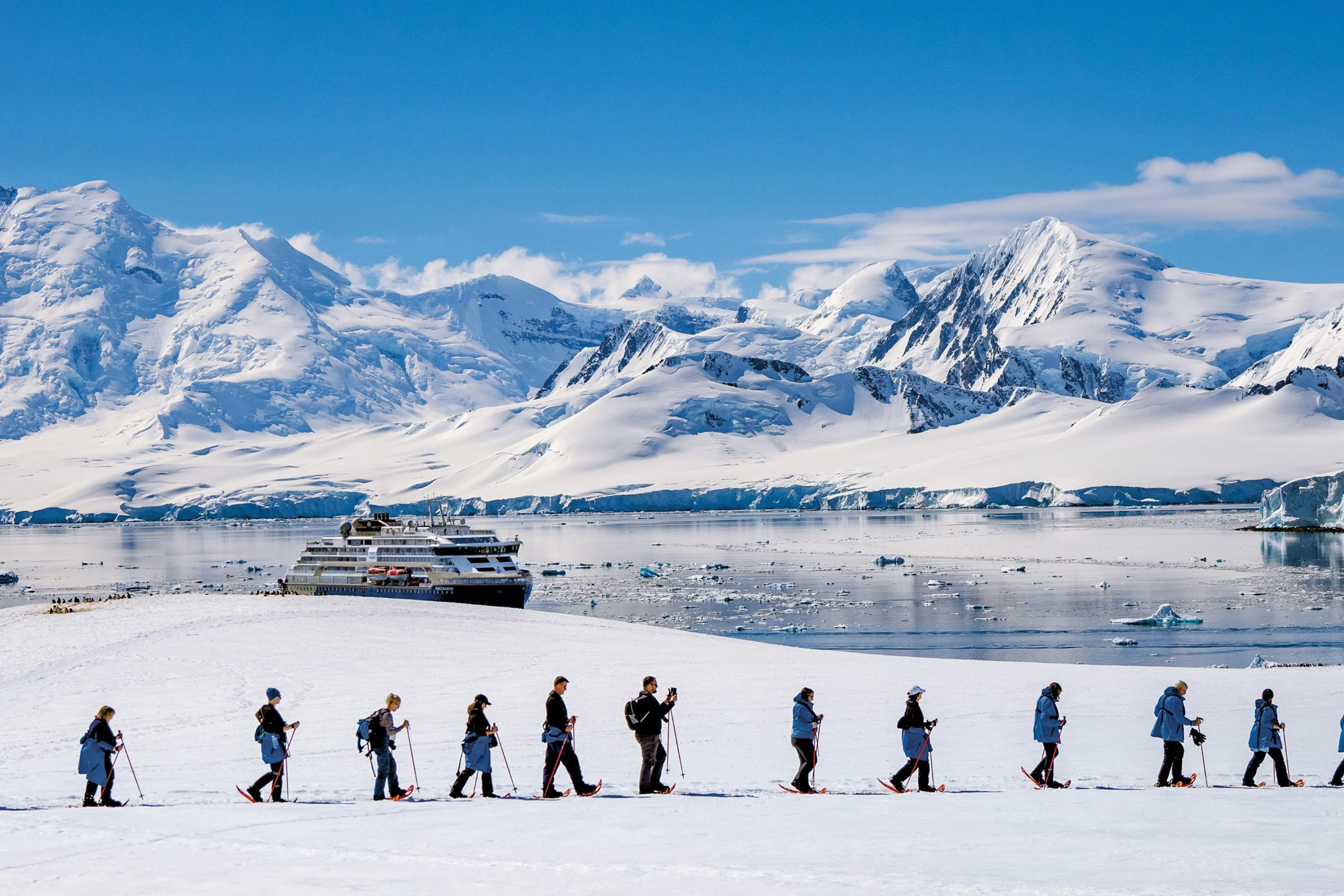 A group of people in winter gear walk in a line across snowy terrain near a calm icy bay, with a large cruise ship and snow-covered mountains in the background under a clear blue sky.