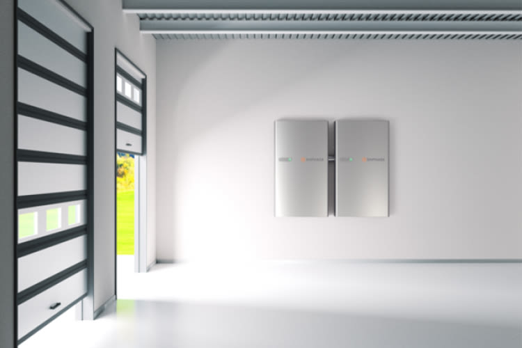 Modern garage interior with two sleek, silver battery storage units called 'Enphase IQ Battery 5P' mounted on a white wall. A garage door is partially open, revealing greenery.