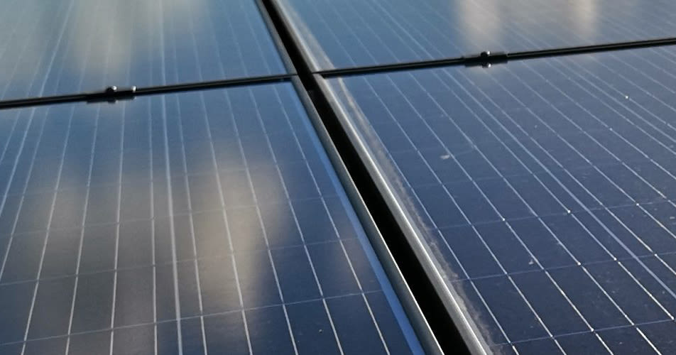 Close-up van zonnepanelen met reflecterende oppervlakken waarin wolkenpatronen zichtbaar zijn. De panelen liggen in een rasteropstelling en geven een gevoel van schone energie en technologie.