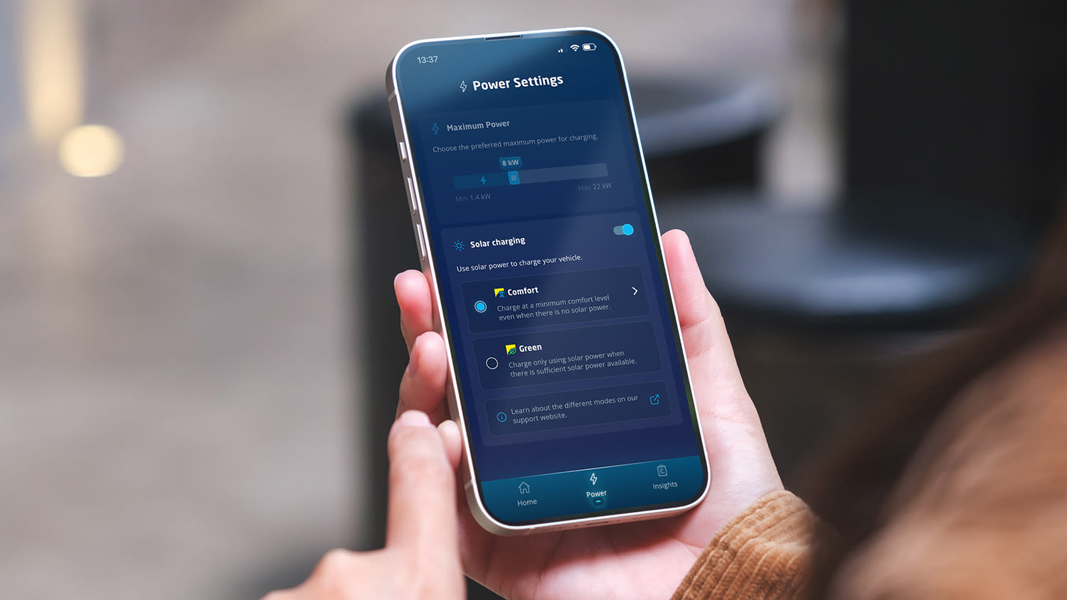 Een persoon houdt een smartphone vast die het scherm "Power Settings" in de Eve Connect-app toont, met nadruk op zonne-opladen en opties voor 'Comfort' en 'Green'. De scène is modern en technologiegericht.