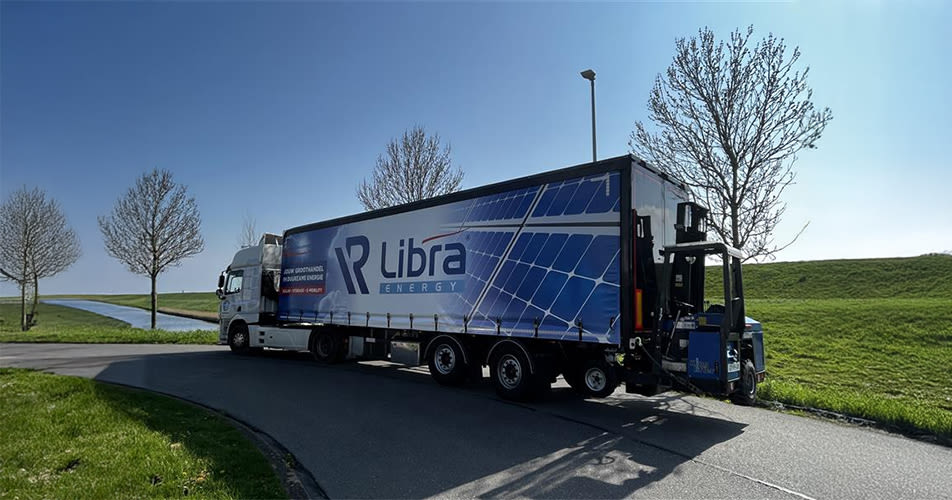A truck with Libra Energy branding showcasing solar panel graphics is driving on a road beside a grassy field and canal under a clear blue sky.