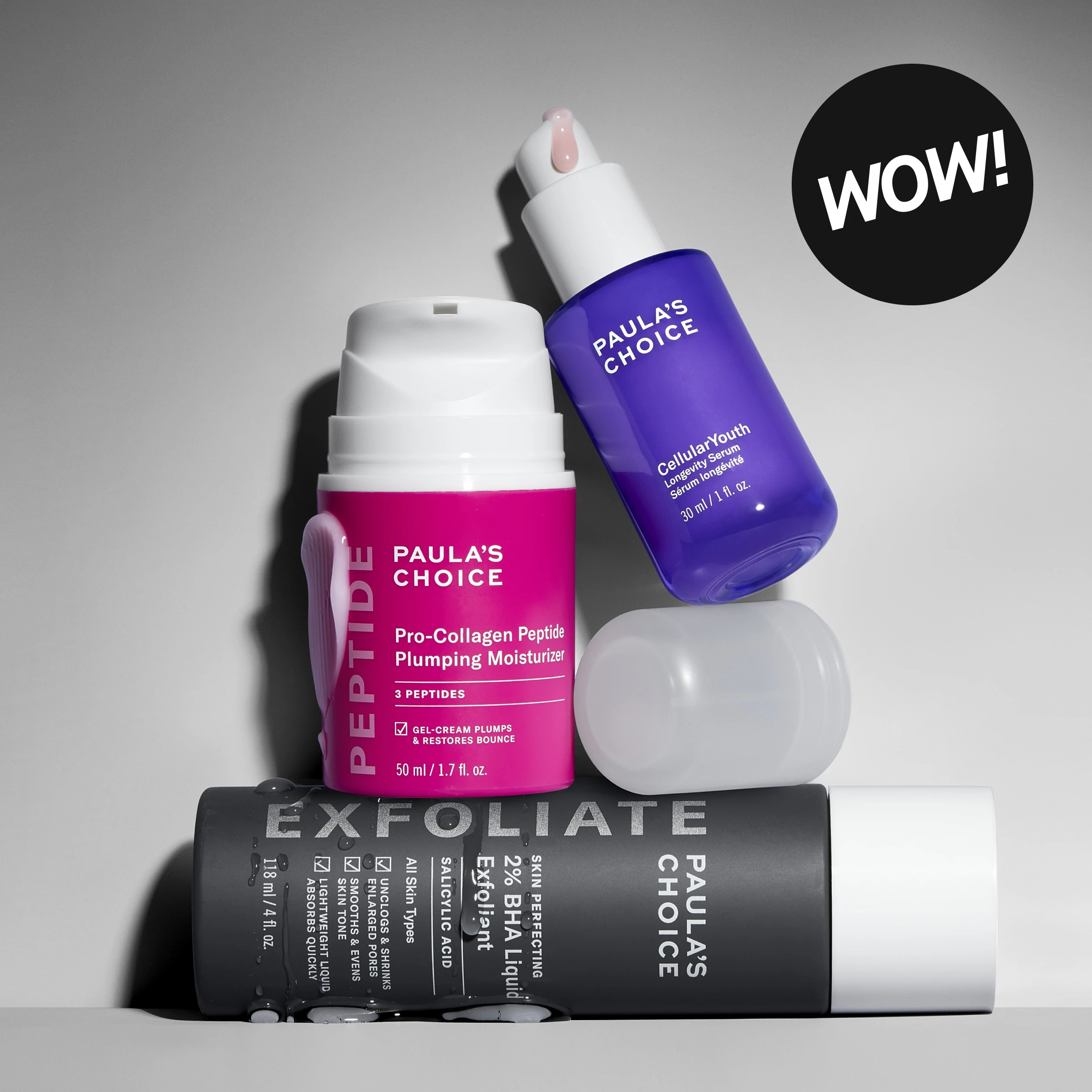 Paula’s Choice-hudpleie stablet mot lys grå bakgrunn med en svart sirkel som inneholder teksten «WOW!». Produktene som vises er Pro-Collagen Peptide Plumping Moisturizer, Cellular Youth Longevity Serum og Skin Perfecting 2% BHA Liquid Exfoliant. Synlig tekst inkluderer «PAULA’S CHOICE», «PEPTIDE», «Pro-Collagen Peptide Plumping Moisturizer», «3 PEPTIDES», «Cellular Youth Longevity Serum», «EXFOLIATE», «2% BHA Liquid Exfoliant», «SALICYLIC ACID» og «WOW!».