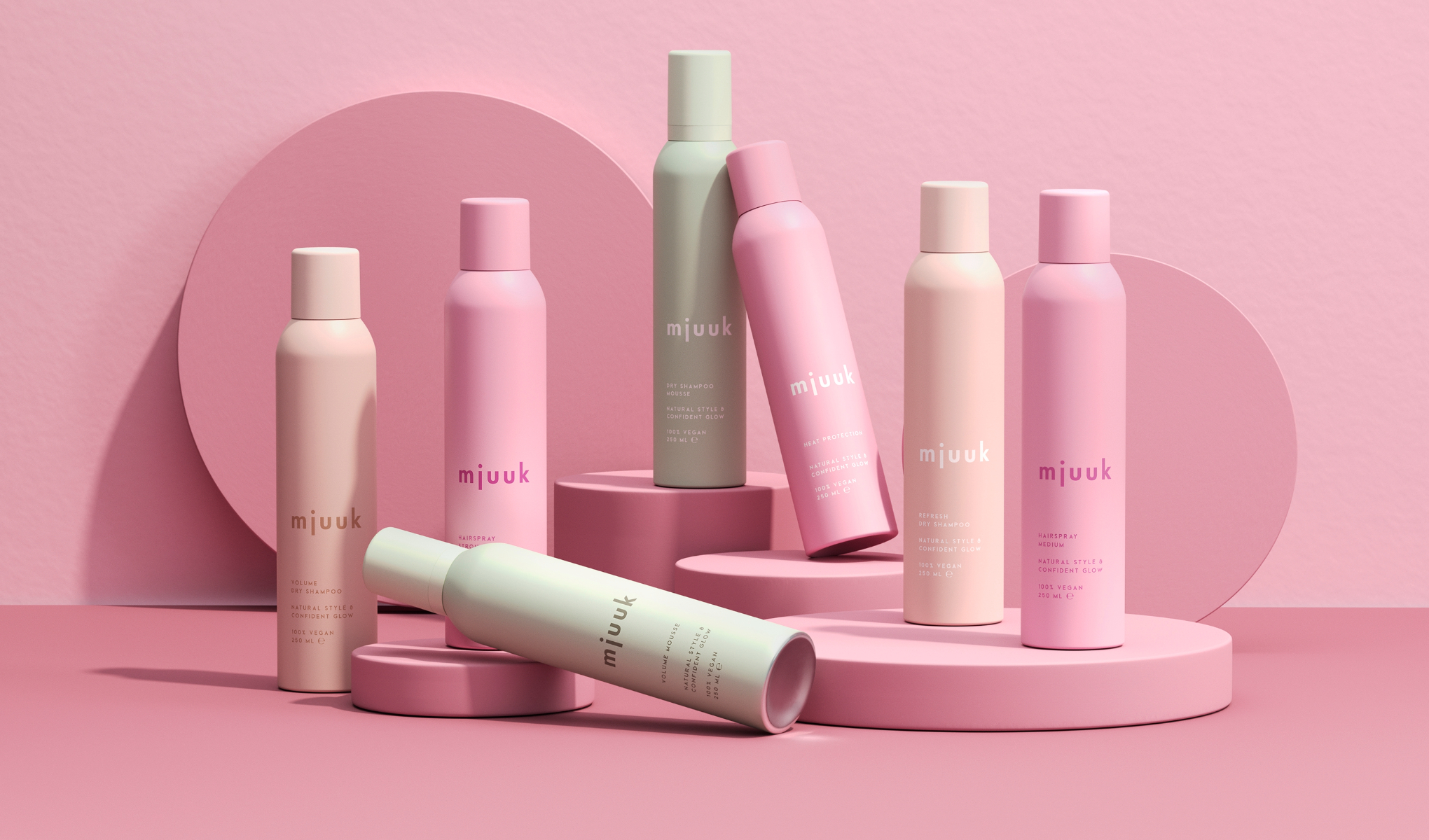 Useita vaaleanpunaisia ja pastellinvärisiä hiustuotteita on aseteltu tyylikkäästi vaaleanpunaiselle taustalle. Tuotteet ovat merkkiä "mjuuk" ja sisältävät muun muassa kuivashampoota ja hiuslakkaa.
