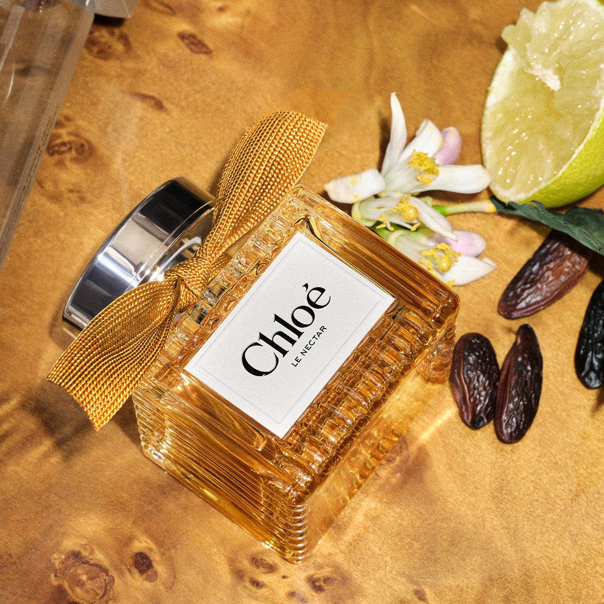 Chloé Le Nectar parfyme i en gyllen glassflaske med gullfarget sløyfe, plassert ved siden av en skåret lime, hvite blomster og mørke frø. Teksten «Chloé» og «LE NECTAR» vises på parfymeflasken.
