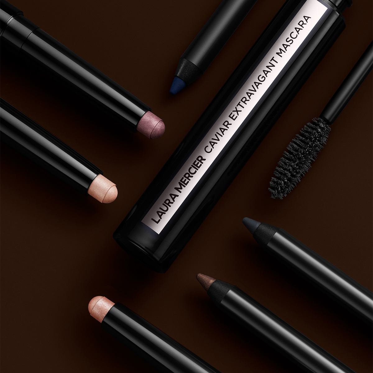 Laura Merciers Caviar Extravagant Mascara omgitt av ulike øyeprodukter, blant annet øyeblyanter og øyenskygger i blyantform, på en mørk bakgrunn.