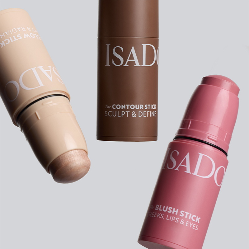 Tre IsaDora makeupsticks i olika nyanser med texten The Contour Stick Sculpt & Define och The Blush Stick Cheeks, Lips & Eyes mot ljus bakgrund. Produkter för contouring och rouge i krämformat.