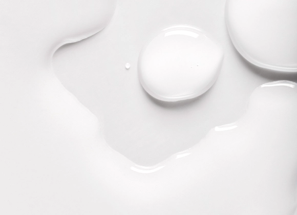 Nærbilde av hvit hudpleie med dråper og myk, glansfull serum- eller lotiontekstur mot hvit bakgrunn. Den kremede konsistensen fremhever et fuktighetsgivende skincare-produkt med lett og silkemyk finish.