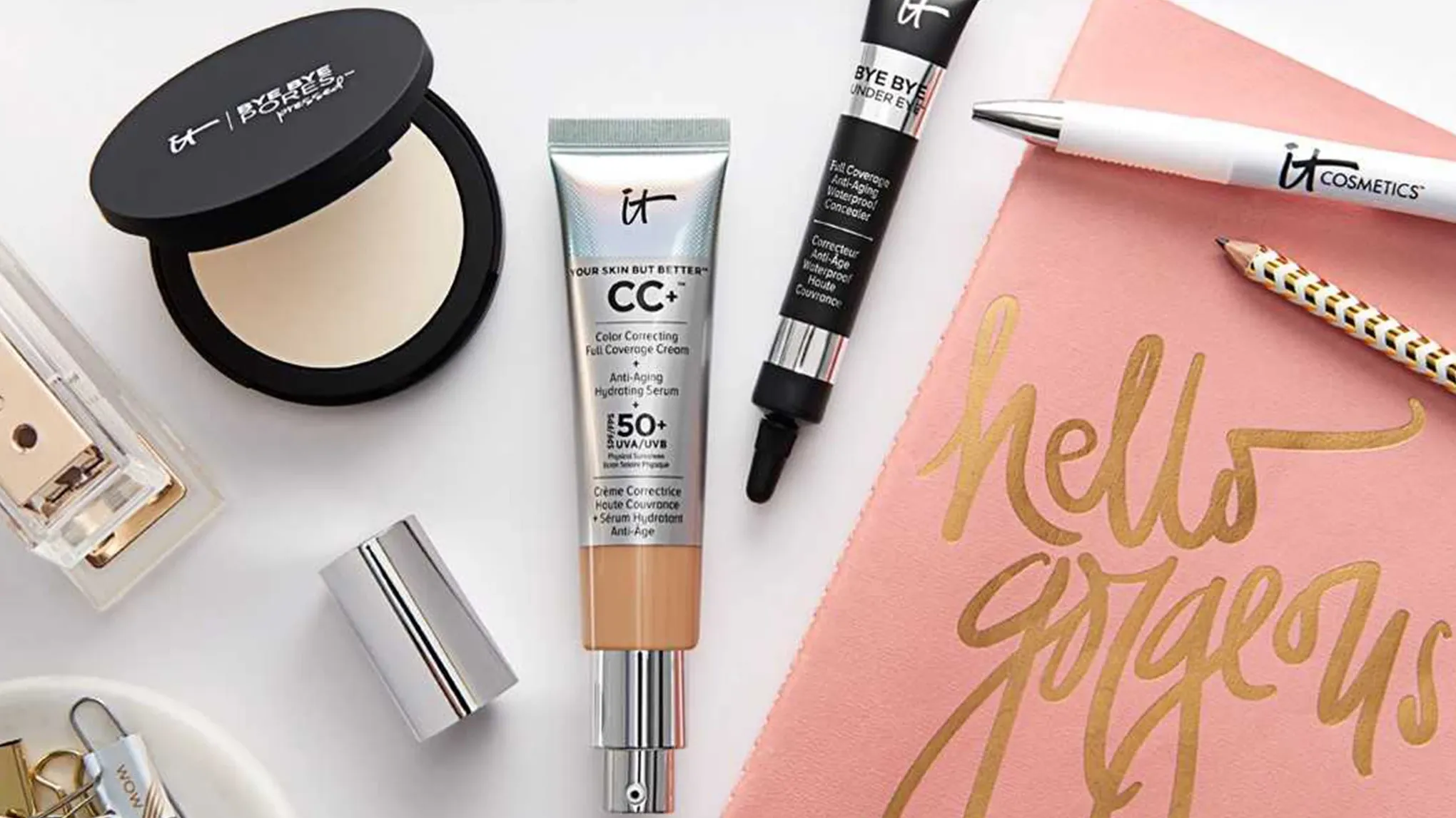 Useita IT Cosmetics -tuotteita aseteltuna valkoiselle alustalle, vaaleanpunaisen liinan kanssa, jossa kultateksti "hello gorgeous".