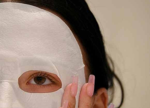 Närbild på en person som har en vit sheetmask i ansiktet. Personen har mörkt hår och rosa naglar. Bakgrunden är beige.