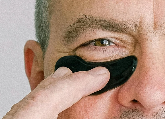 Närbild på en person med en svart ögonmask under ögat som han håller fast med ett finger. Personen är gråhårig och bakgrunden är ljusgrå.