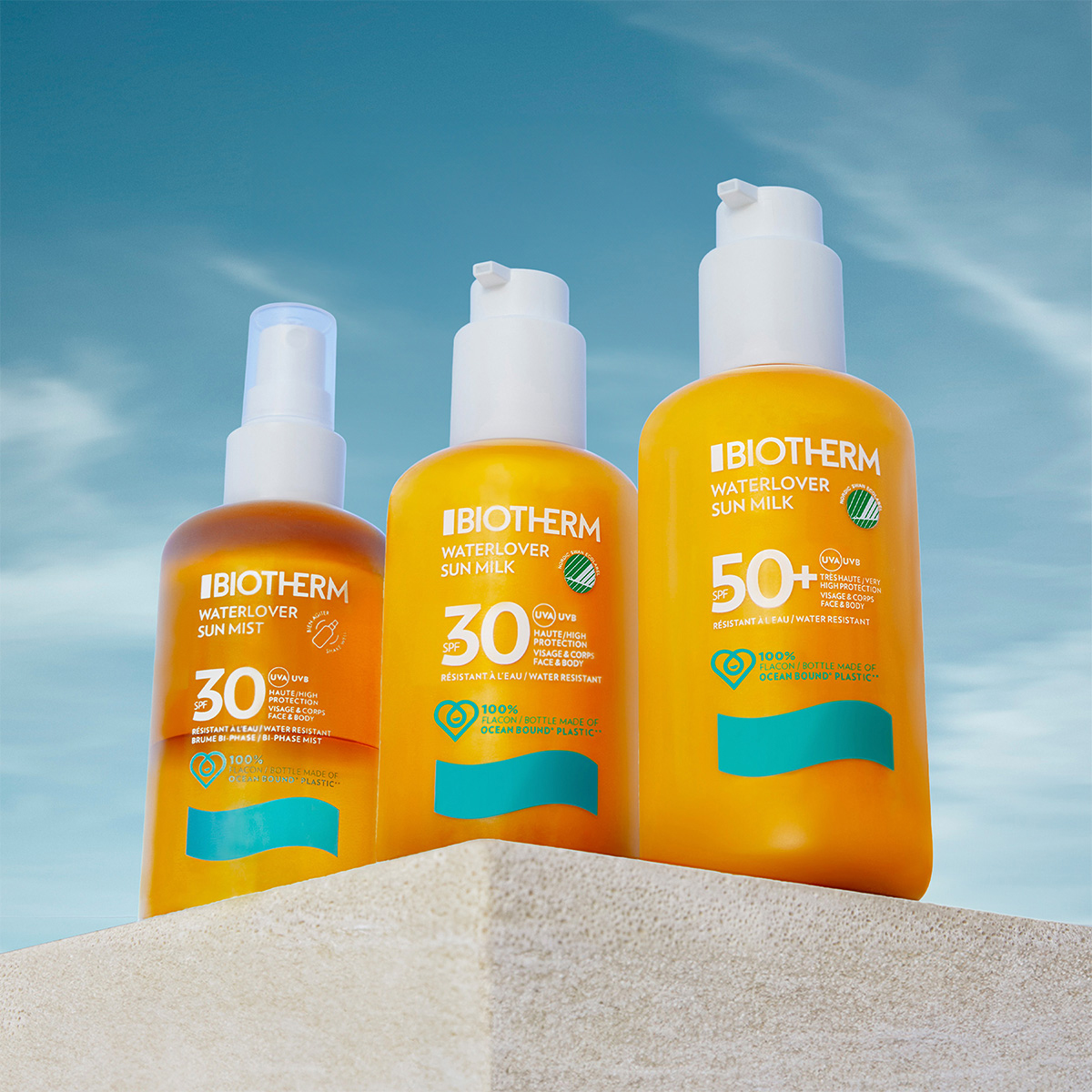 Tre solskyddsprodukter från Biotherm är placerade på en beige plattform mot en blå himmel. Produkterna inkluderar: en brun sprayflaska, en gul pumpflaska och en större gul pumpflaska.
