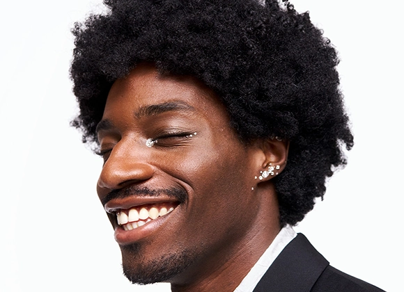 Bild som visar en person med svart lockigt hår och flera örhängen i örat. Personen bär en svart kavaj och en vit skjorta. Bakgrunden är vit.