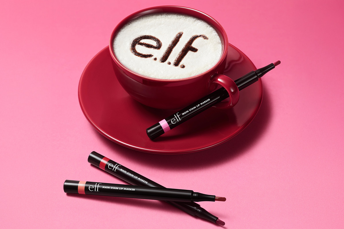 e.l.f. Main Stain Lip Marker -huulivärejä vaaleanpunaisella taustalla punaisen kahvikupin vieressä. Kuvassa teksti: ”NEW!”.