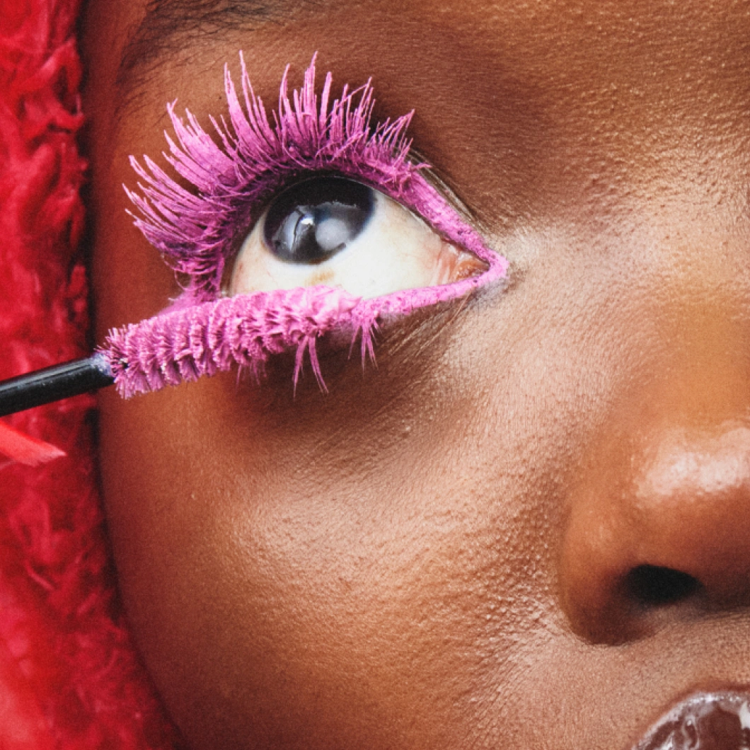 Närbild på ett öga med intensiv rosa mascara på både övre och nedre ögonfransar. En mascaraborste med rosa produkt hålls mot de nedre fransarna.