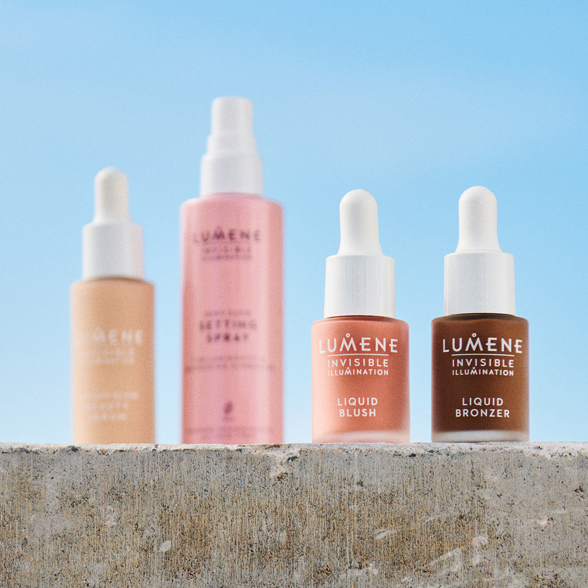 Fyra Lumene hudvårdsprodukter på en betongyta utomhus. Foundation, setting spray, flytande rouge och bronzer.