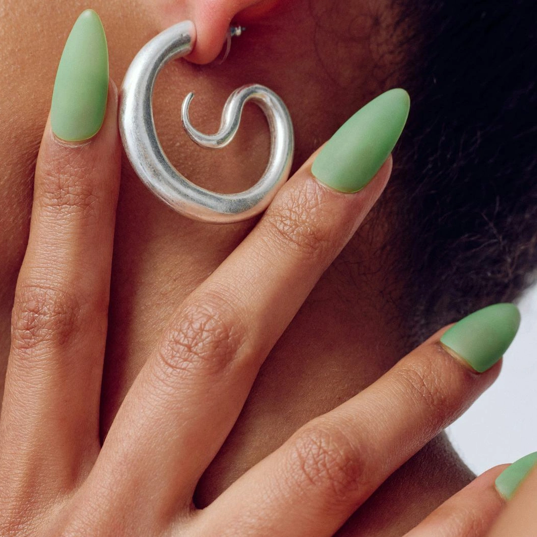 Närbild på en hand med långa, mandelformade naglar målade i matt ljusgrönt nagellack. Fingrarna hålls intill en stor silverfärgad spiralformad öronring.